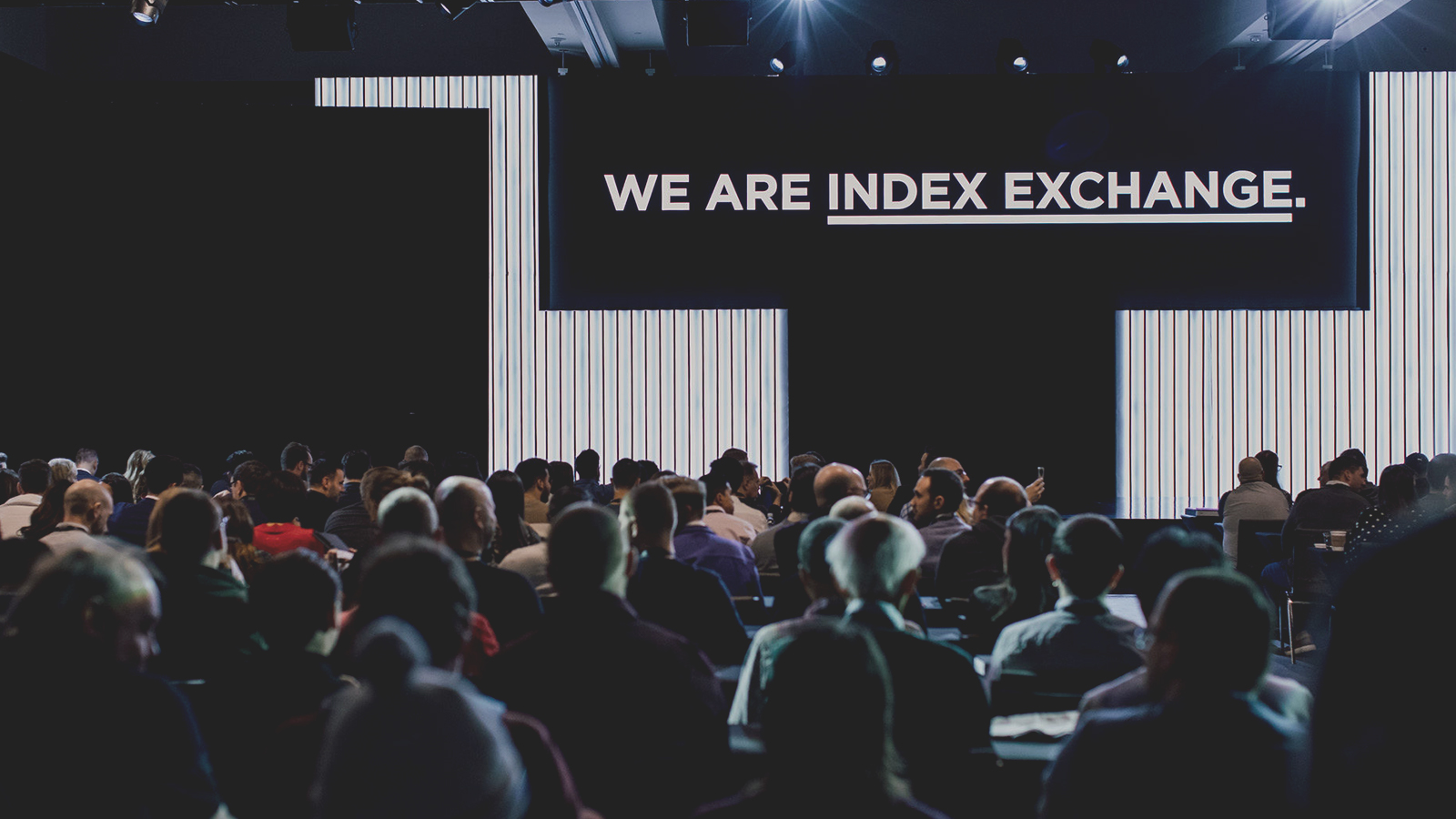 Imagen que representa a personas sentadas en un centro de conferencias mirando un escenario que tiene una pantalla que dice somos index exchange