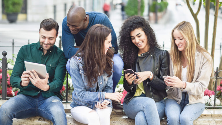 Groupe de jeunes personnes assises sur un rebord en pierre dans la rue, discutant en regardant des contenus sur leur téléphones mobiles et tablettes